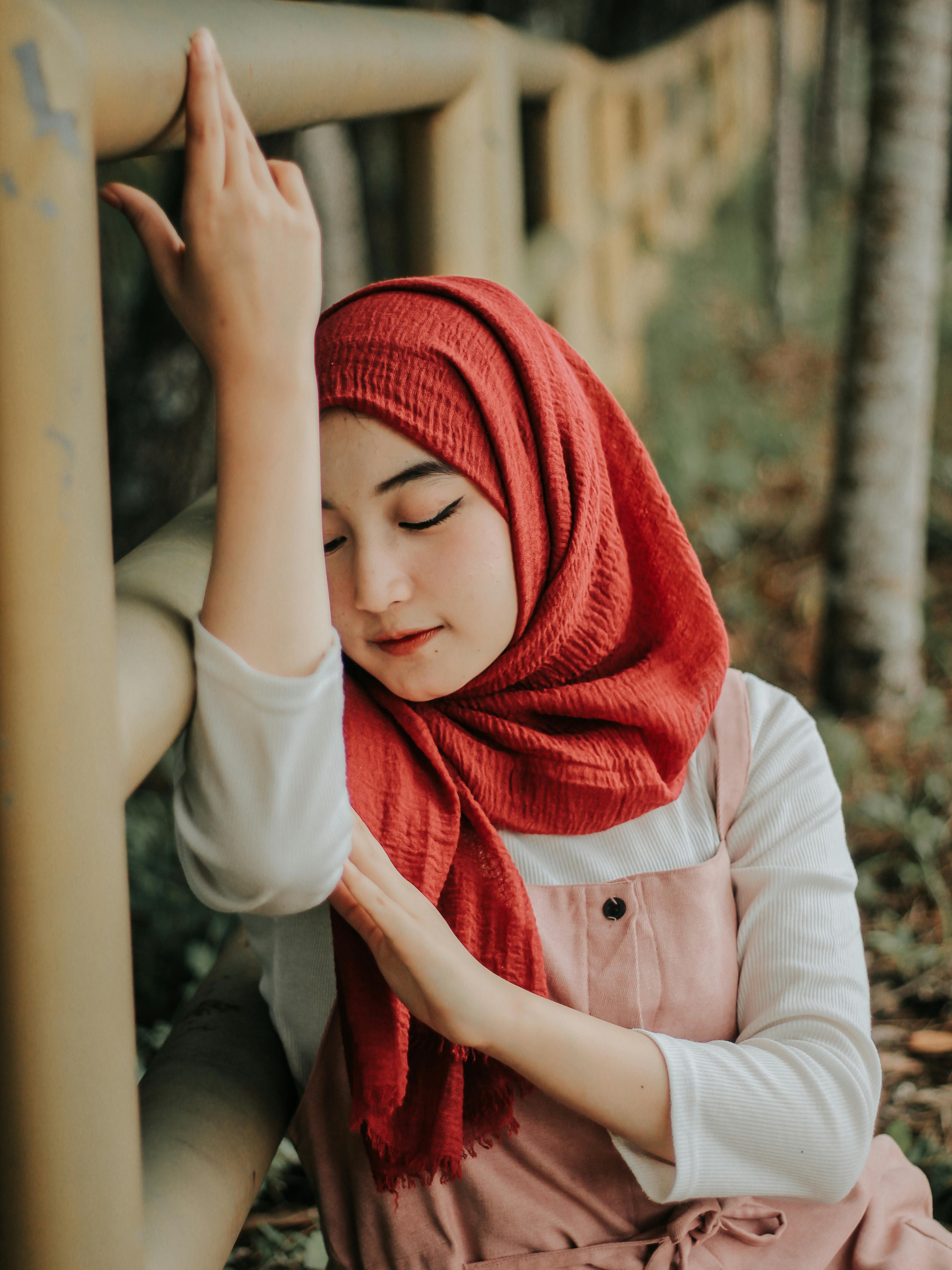 Peaceful young woman wearing a hijab resting thoughtfully against a fence outdoors.