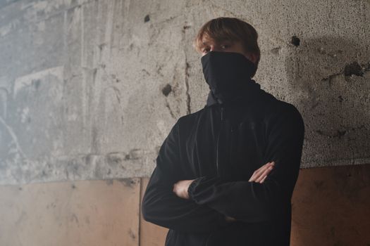 A young man in a black jacket faces the camera, wearing a mask in a rustic indoor setting, embodying the new normal.