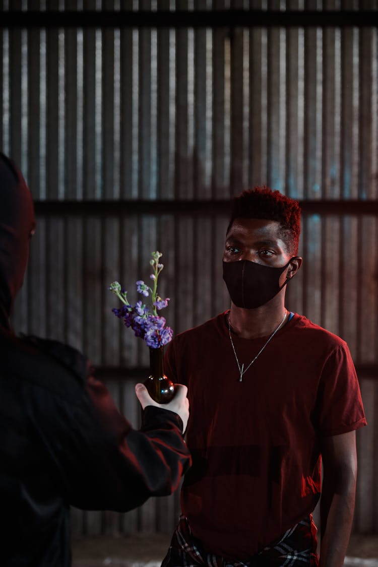 A Person In Black Jacket Handing A Vase With Flowers To The Man Wearing Red Shirt