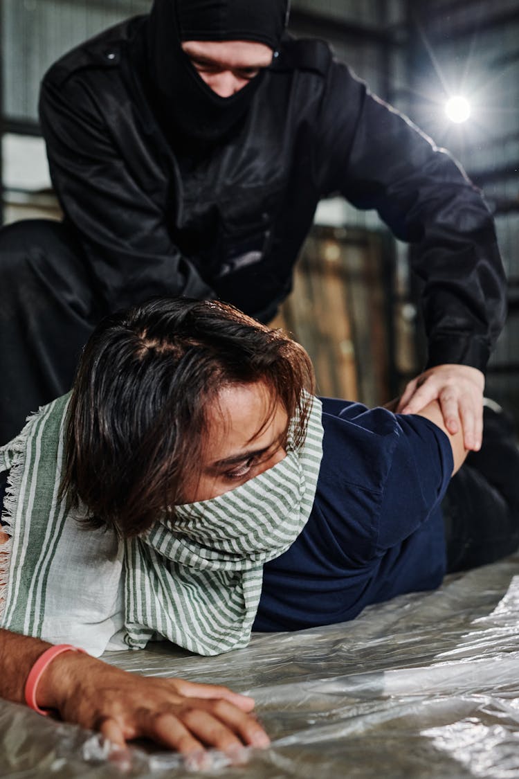 A Man In Black Balaclava Twisting The Arm Of The Person Lying On The Ground