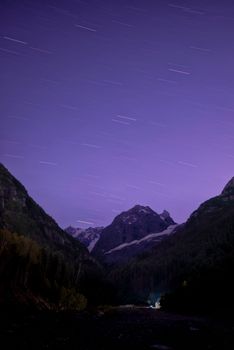 Long exposure of star trails over mountains at night, showcasing a vibrant purple sky.
