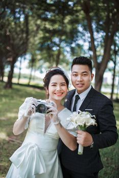 Smiling couple dressed stylishly in wedding attire holding camera and bouquet outdoors.