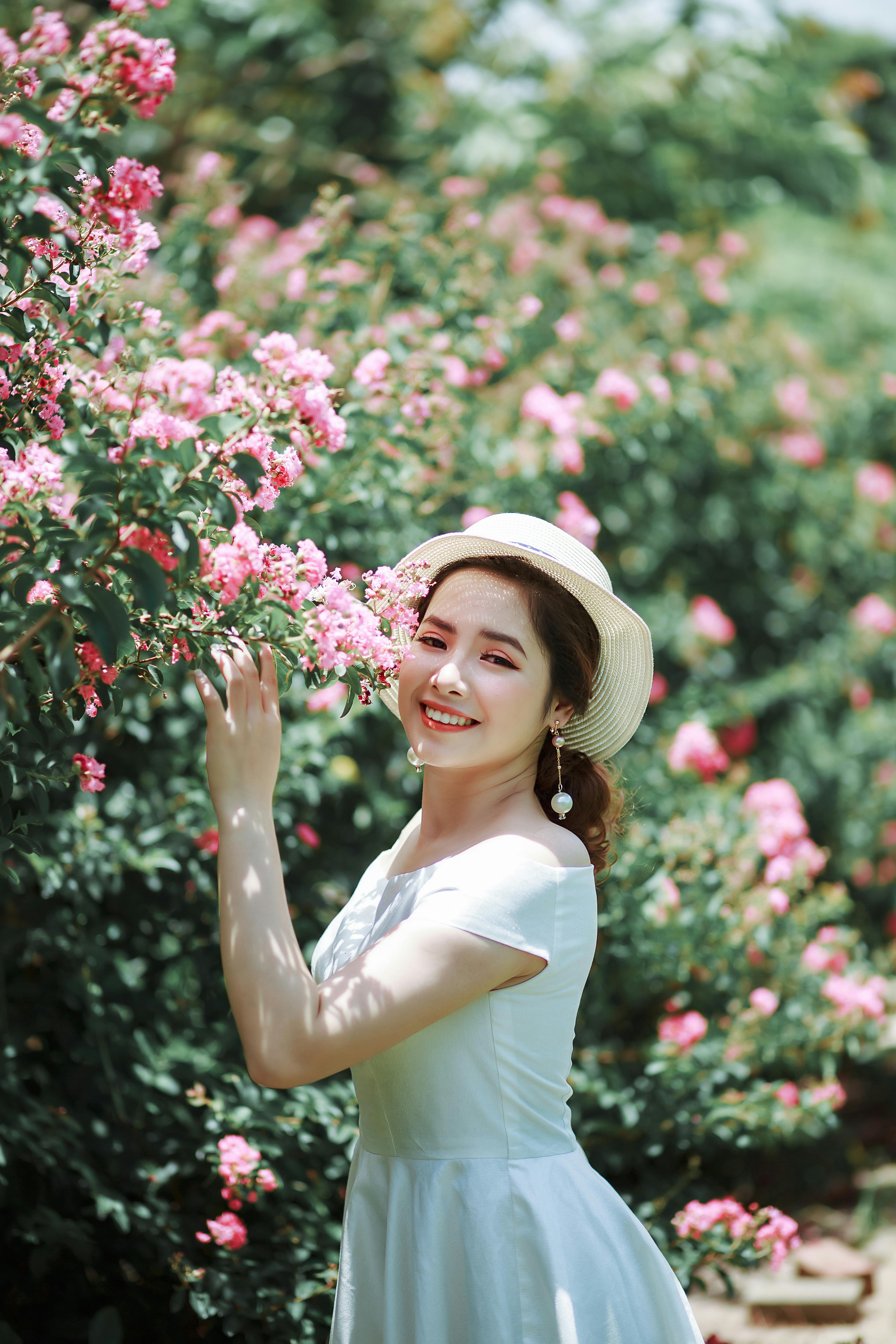 A Woman in White Dress Touching the Pink Flowers while Smiling at the  Camera  Free Stock Photo
