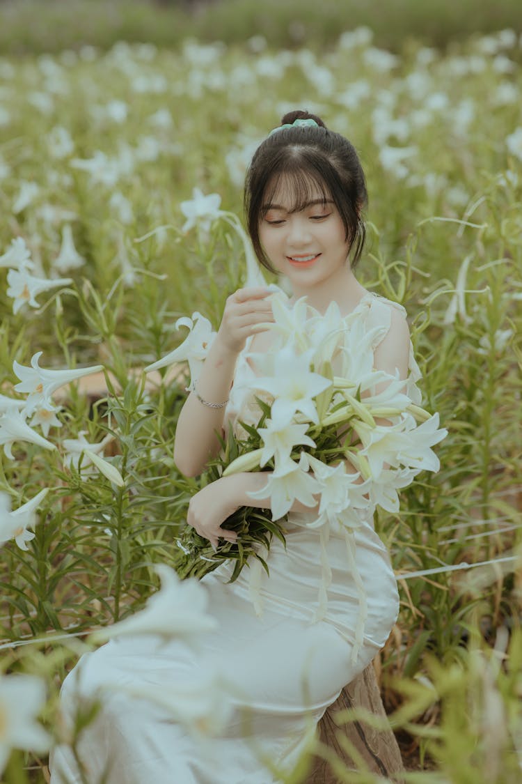 Young Woman In A White Dress Sitting On A Field Holding A Bunch Of White Lilies 