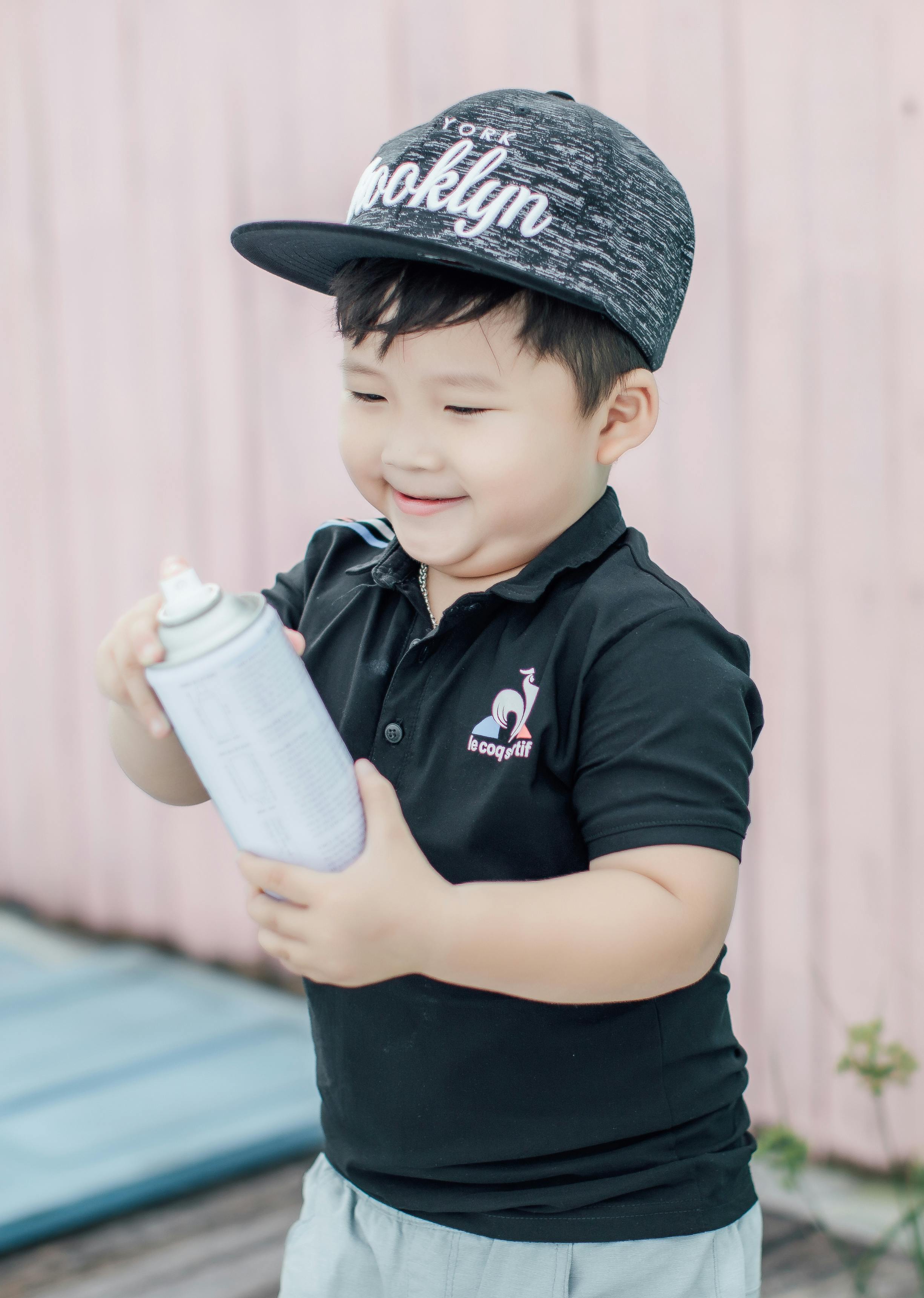 An adorable Asian boy in a cap joyfully holding a spray paint can outdoors.