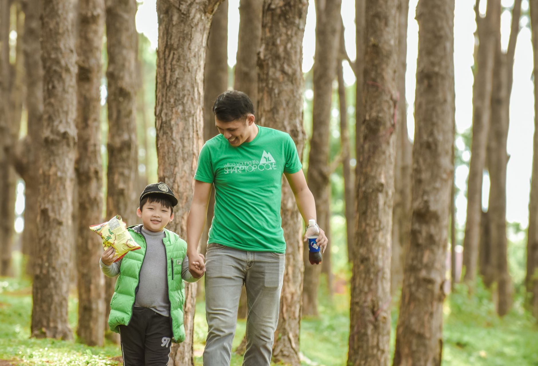 A smiling father and his son walking hand in hand in a sunlit forest, bonding and enjoying nature.