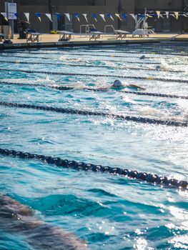 Swimmers competing in a sunny, outdoor swimming pool race with lanes and water splashes visible.