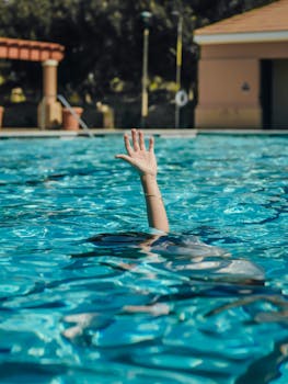 A person's hand raised above the water in a clear swimming pool, suggesting swimming or asking for help.