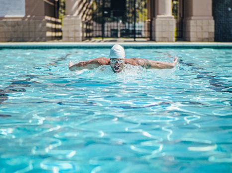 A man wearing goggles and a swimming cap performing a butterfly stroke in an outdoor swimming pool.