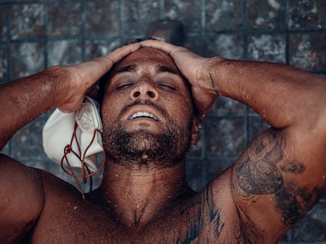 Close-up of a shirtless man with tattoos enjoying a refreshing moment under water, hands on head, eyes closed.