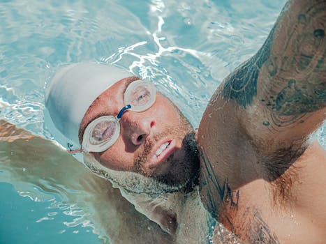 Close-up of a male swimmer with tattoos practicing in a pool, wearing goggles and cap.