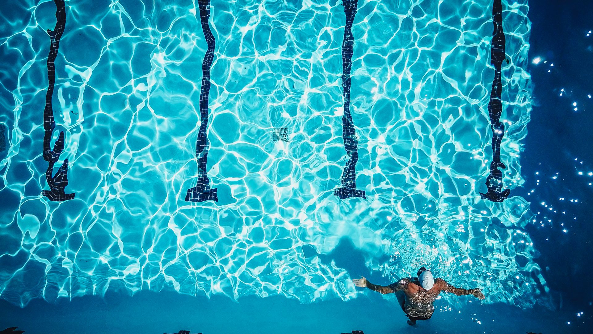 A lone swimmer doing laps in a vibrant turquoise pool, captured from above on a sunny day.