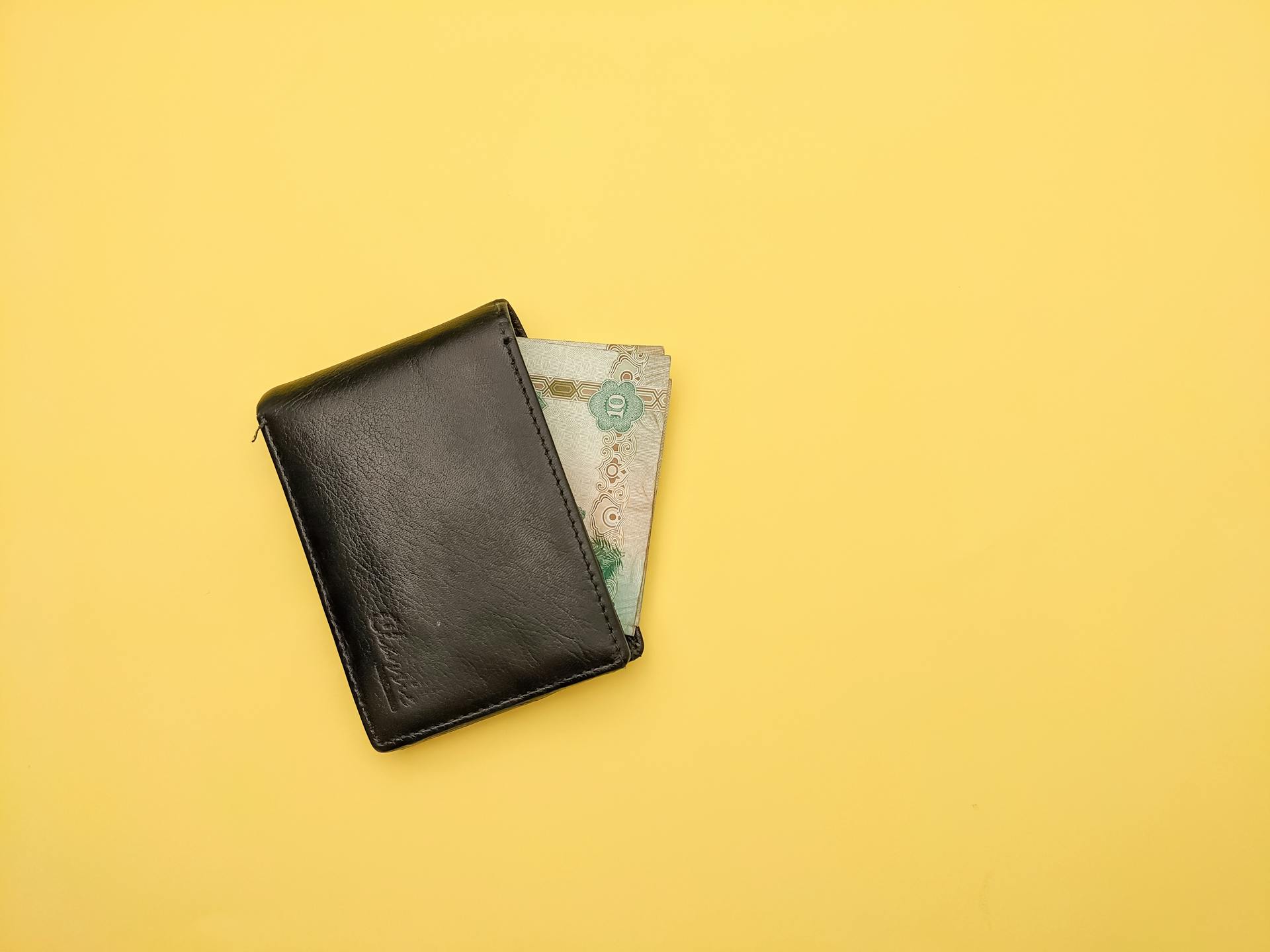 A black leather wallet partially open with visible paper currency on a bright yellow background.