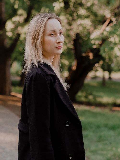 Free A woman in a black coat stands outdoors in a park with lush greenery, during daytime. Stock Photo