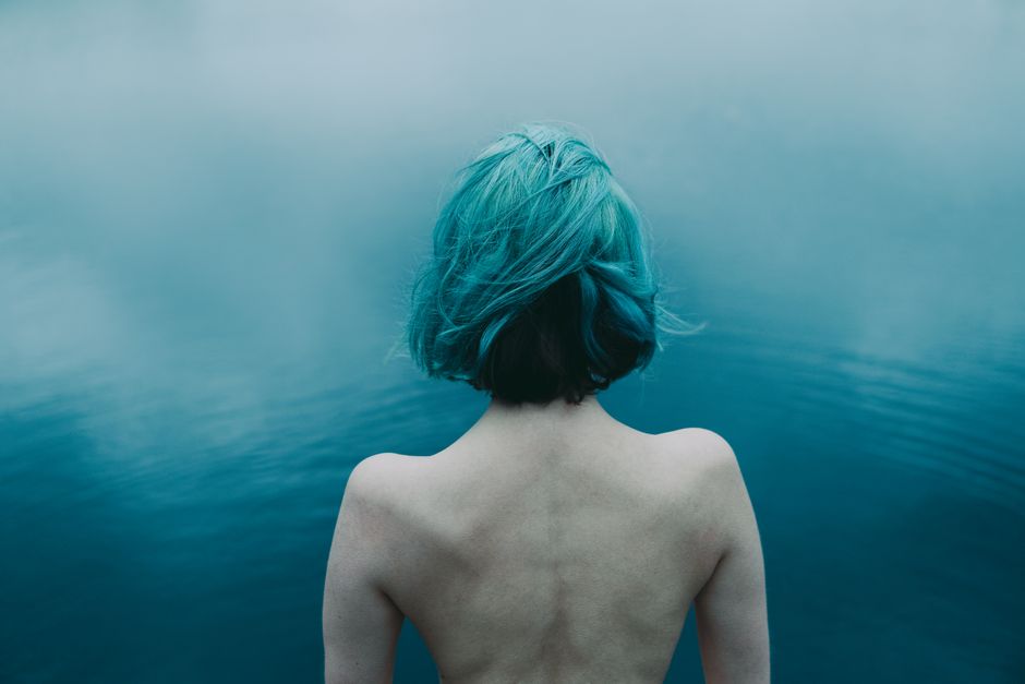 A creative back view of a woman with teal hair by a serene lake, conveying calm and mystery.