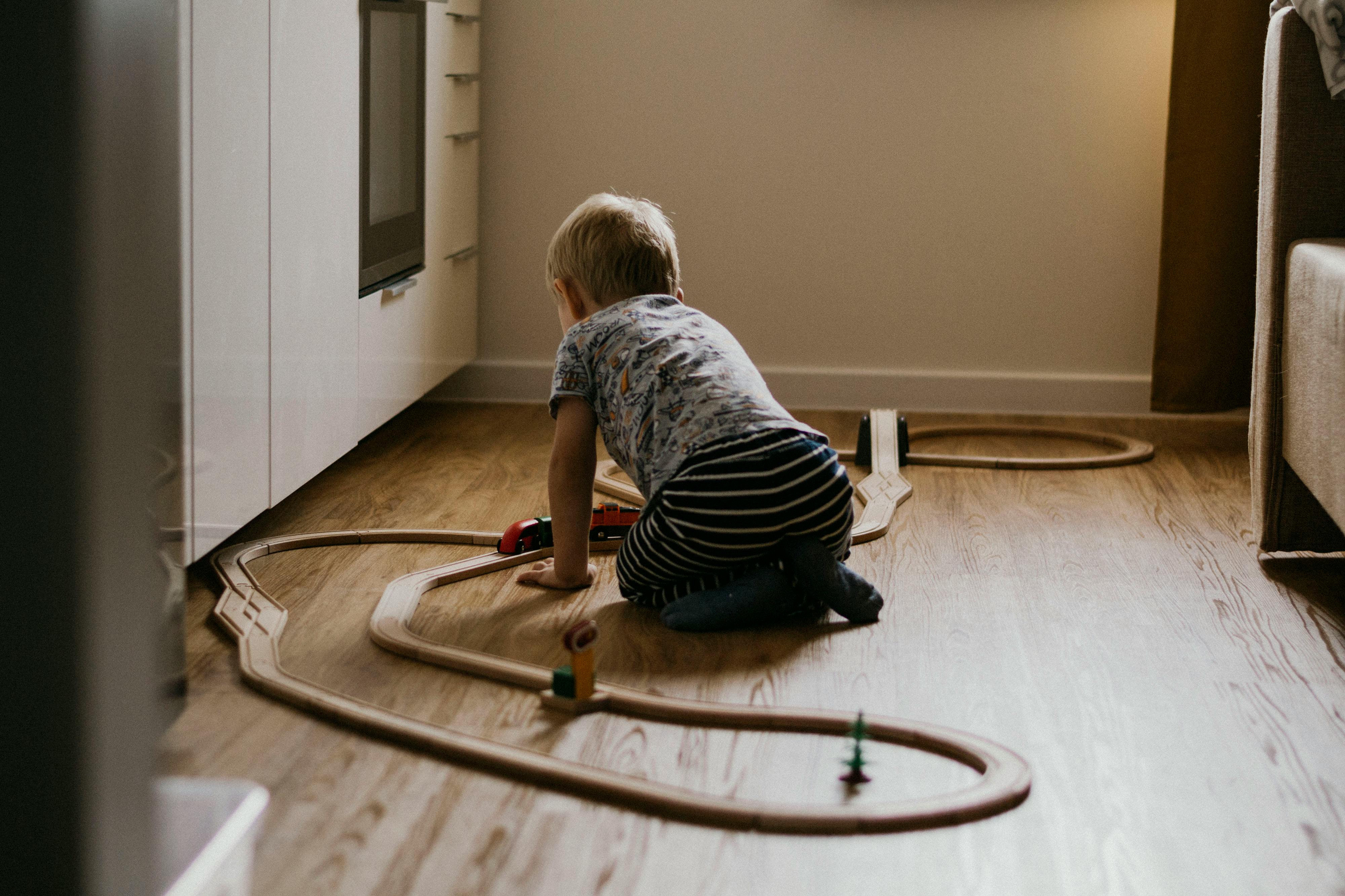 A young child sits on the floor, engaged with a wooden train set in a cozy indoor setting.