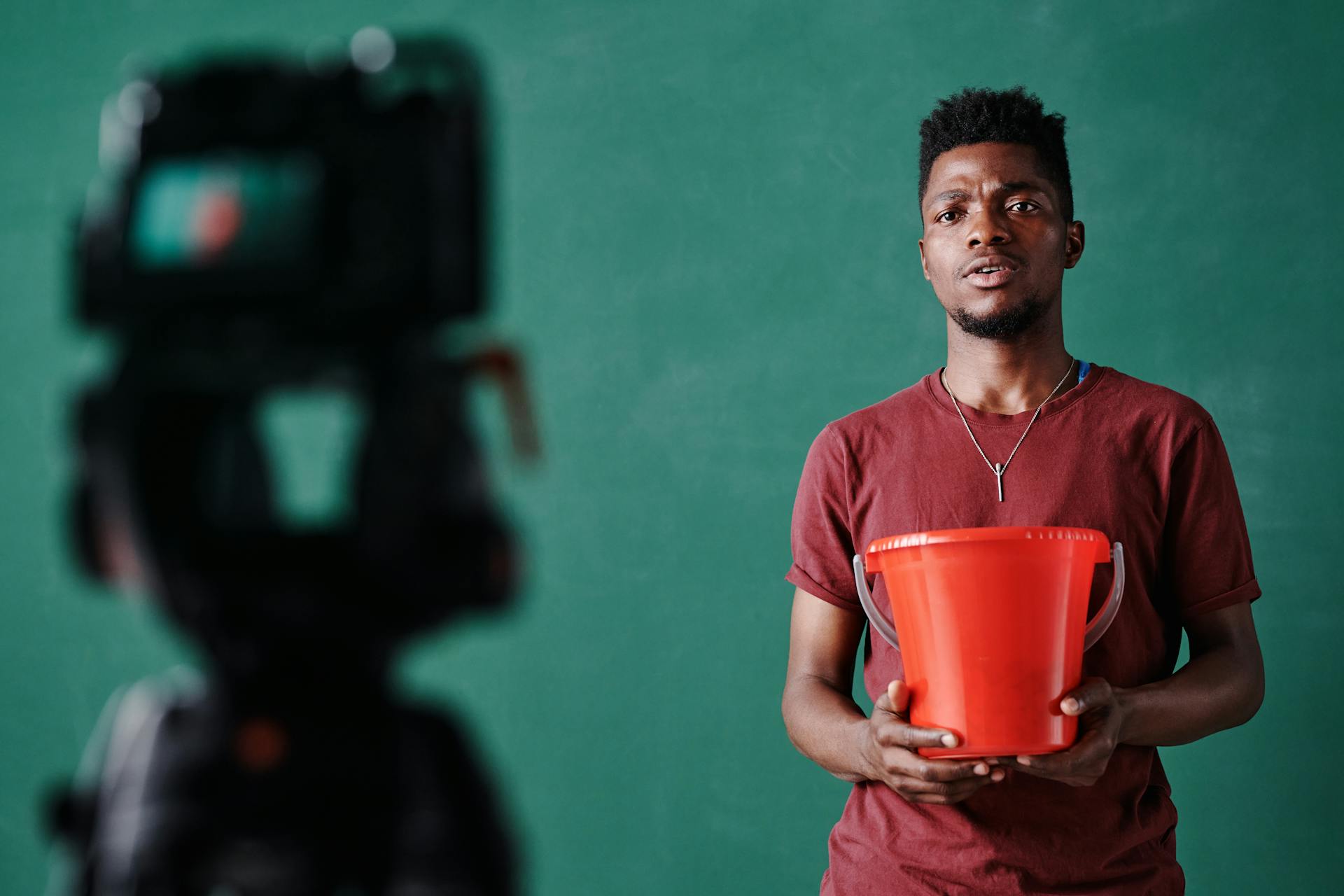 Black man holding a red bucket in front of a camera setup with green backdrop, preparing for a vlog.