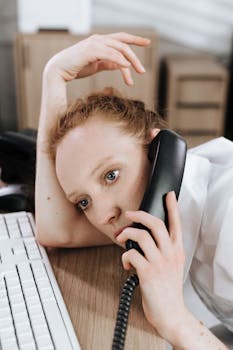 A tired call center agent leans on a desk while holding a telephone handset, portraying workplace fatigue.
