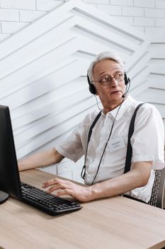 Senior man with headset assisting customers at call center using a desktop computer.