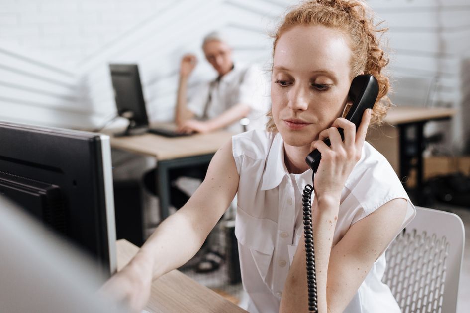 Une employée en chemise blanche répond à un téléphone filaire à son bureau, les yeux sur son écran, dans un open space clair.