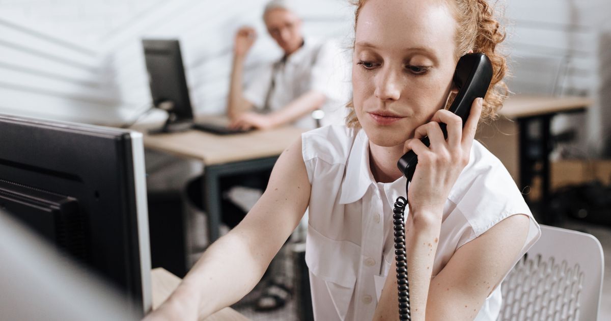 Une employée en chemise blanche répond à un téléphone filaire à son bureau, les yeux sur son écran, dans un open space clair.