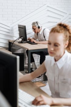 An elderly man wearing a headset works at a computer in a modern office setting.