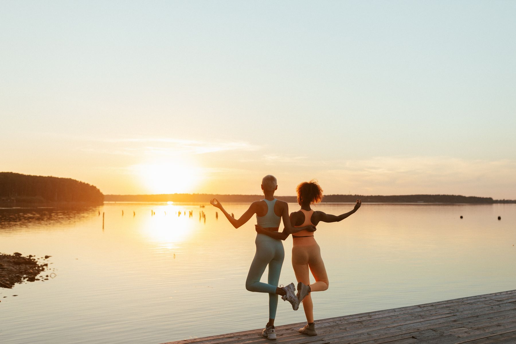 Two women balancing side by side in tree pose on a lakeside boardwalk, facing a low golden sun over the water