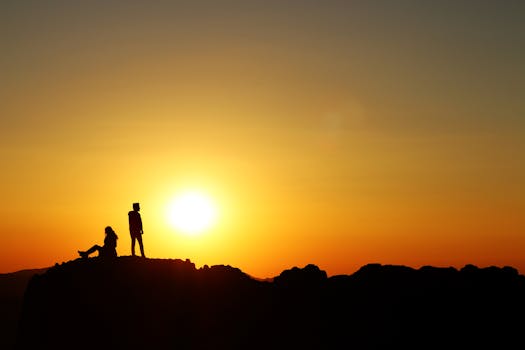 Silhouette of a person standing and another sitting on a hill at sunset in Türkiye, capturing the golden glow of the evening sky.