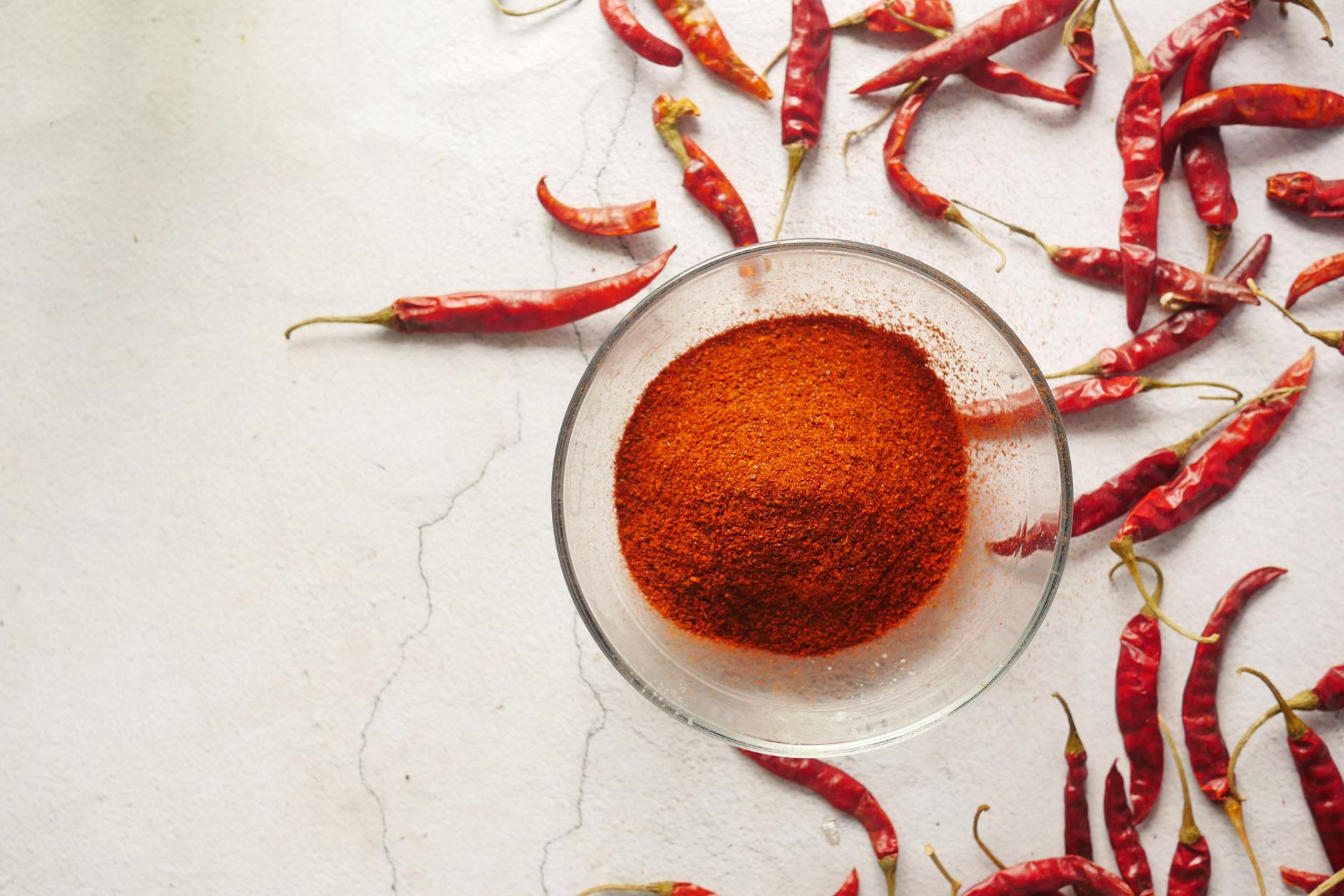 Cayenne Pepper Powder In A Small Bowl With Fresh Cayenne Peppers And A Glass Of Lemon Water On A Kitchen Countertop
