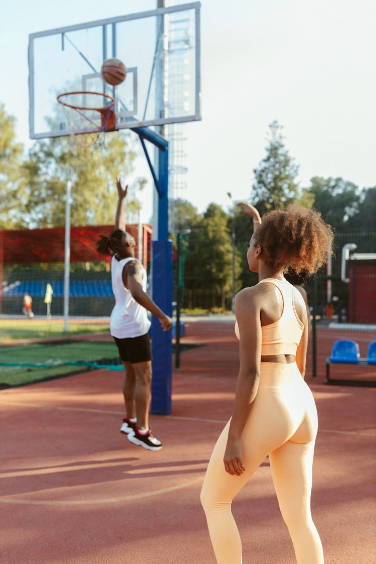 Woman In Beige Activewear Standing In A Basketball Court  Near A Man Shooting The Ball 