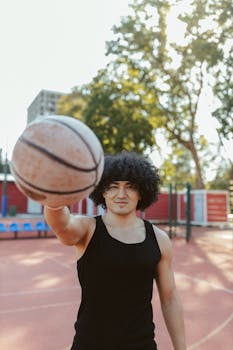 A young man with afro hair holding a basketball on an outdoor court during the day.
