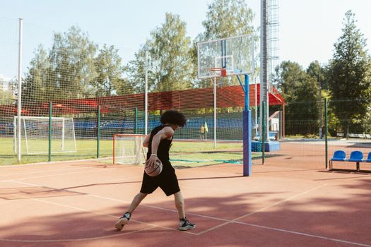 A young man dribbles a basketball on an outdoor court with surrounding greenery.