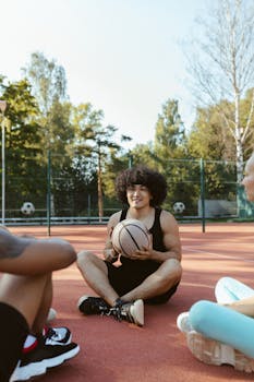 Smiling young man sits with friends holding a basketball on an outdoor court.