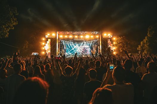 Large crowd enjoying a spectacular live outdoor concert at night with stage lighting effects.