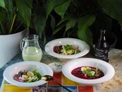 A sophisticated dining table setup featuring beetroot soup, salads, a bottle of drink, and a pitcher amidst lush greenery.