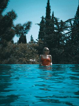 Woman in a pool wearing orange swimwear, surrounded by lush trees, exuding serenity.