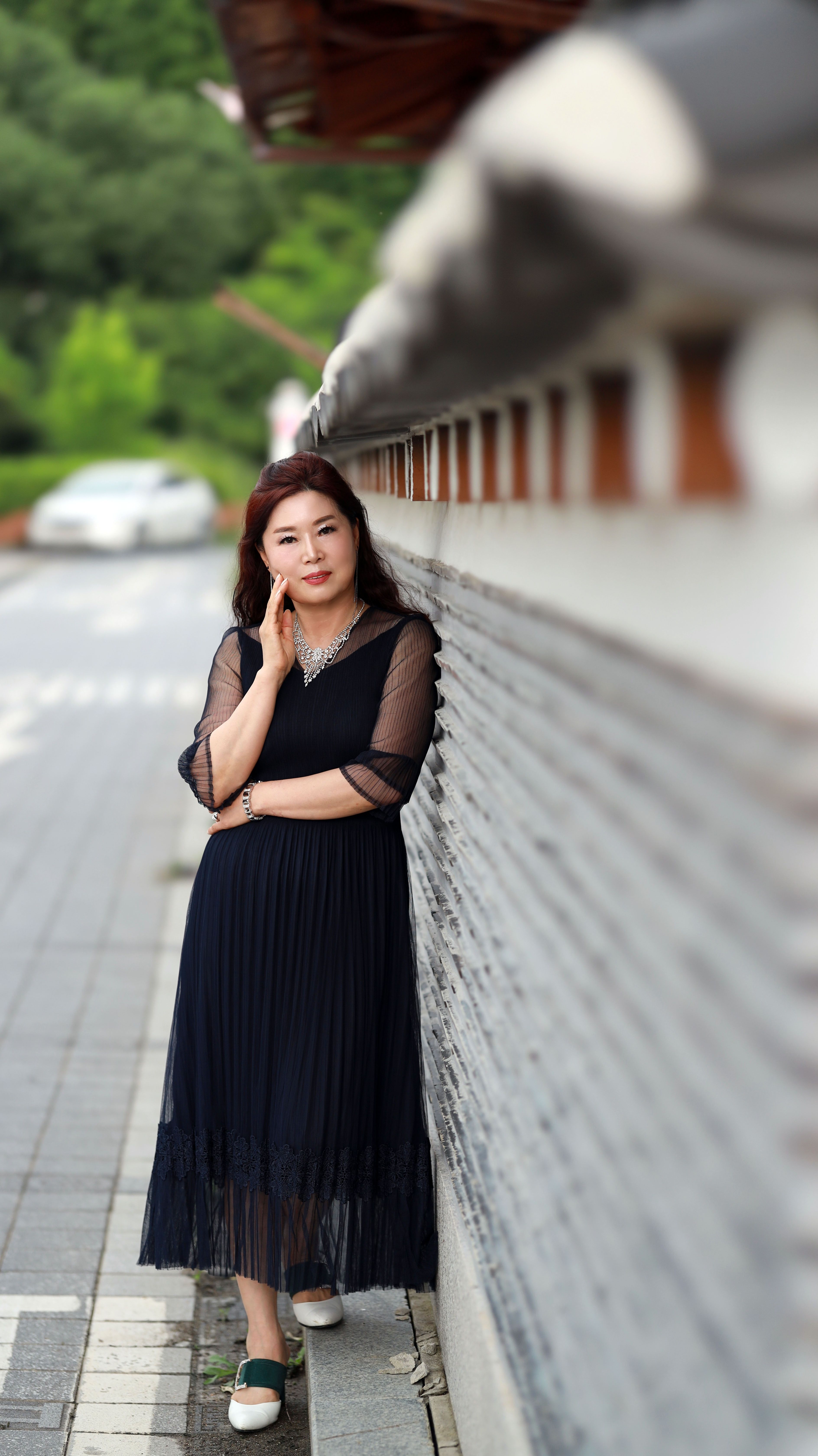 Elegant woman in black dress leaning against a wall on a quiet street.