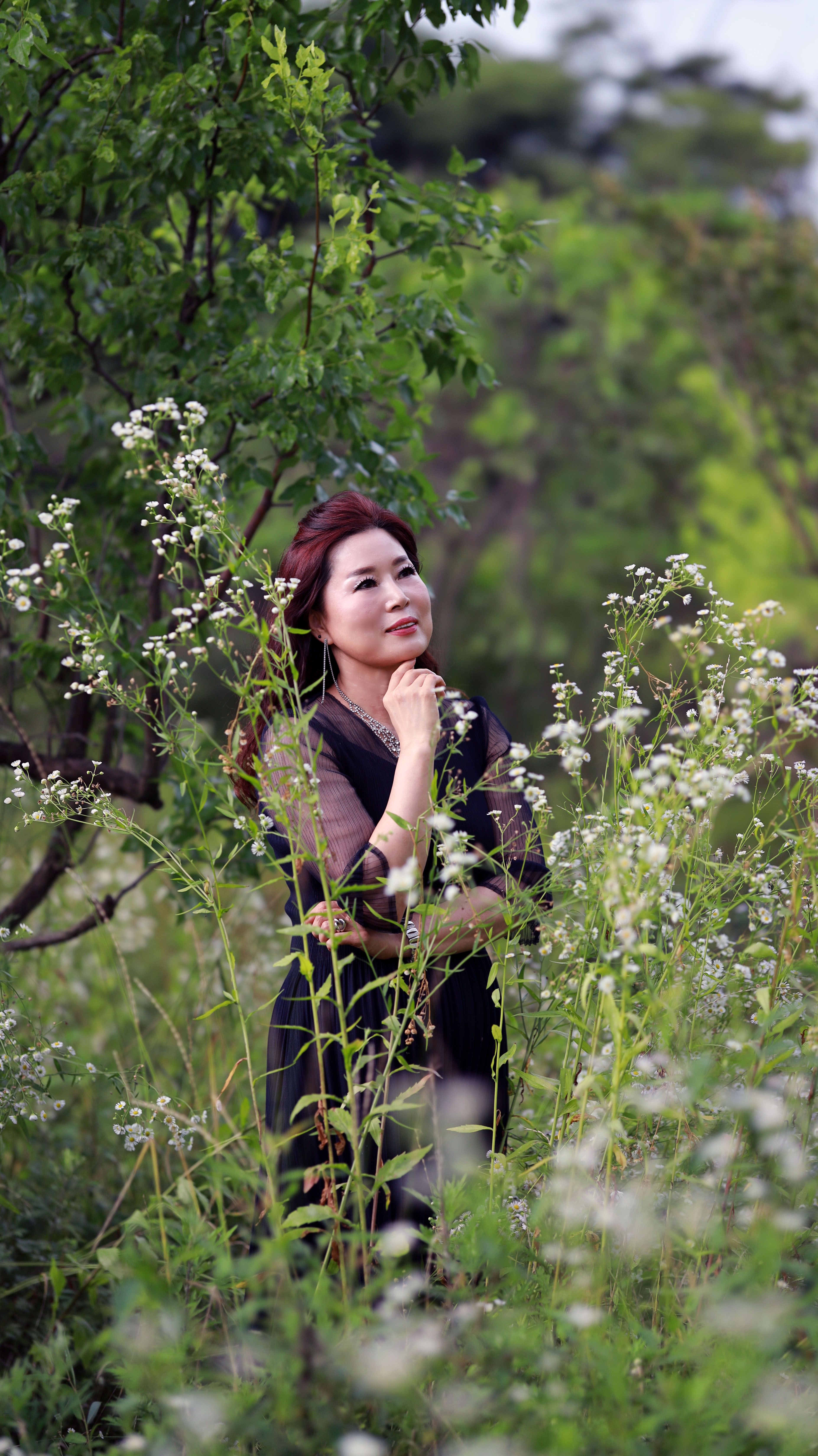 A thoughtful woman in a black dress standing amidst lush greenery, embodying elegance and tranquility.