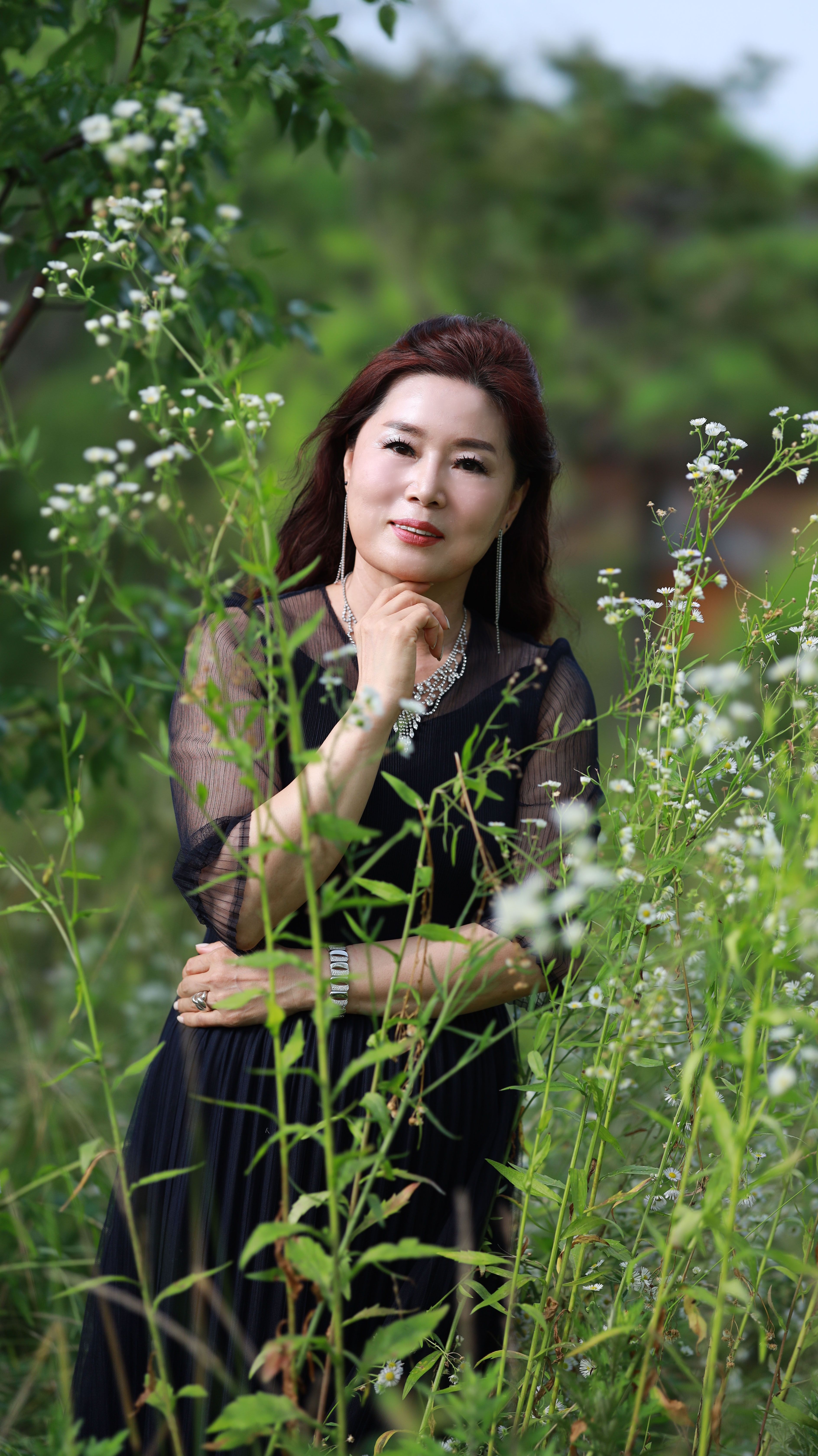 A woman in a black dress poses gracefully among tall plants in a serene outdoor setting.