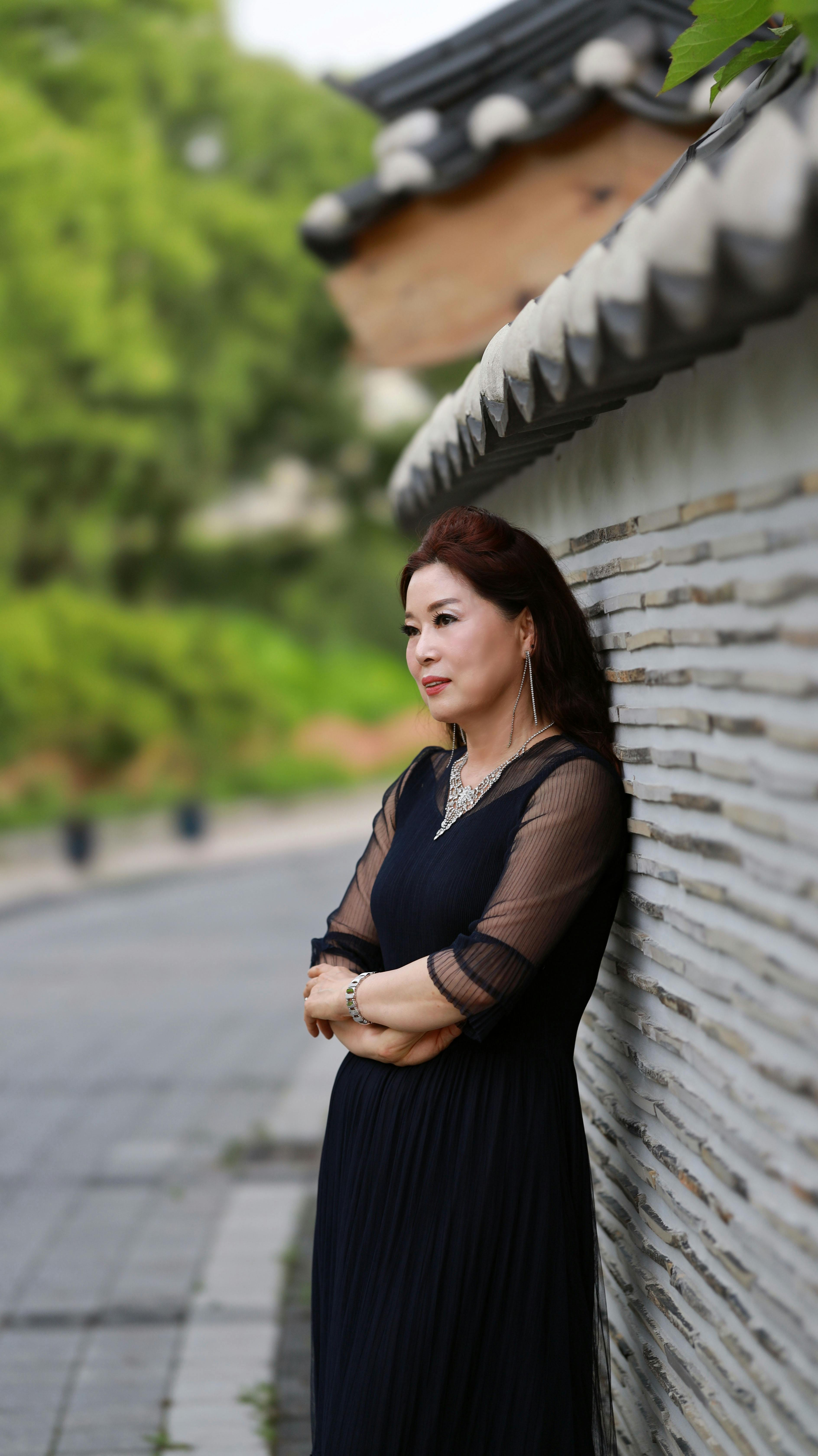 Elegant woman in black dress posing by traditional wall outdoors, exuding grace and fashion.