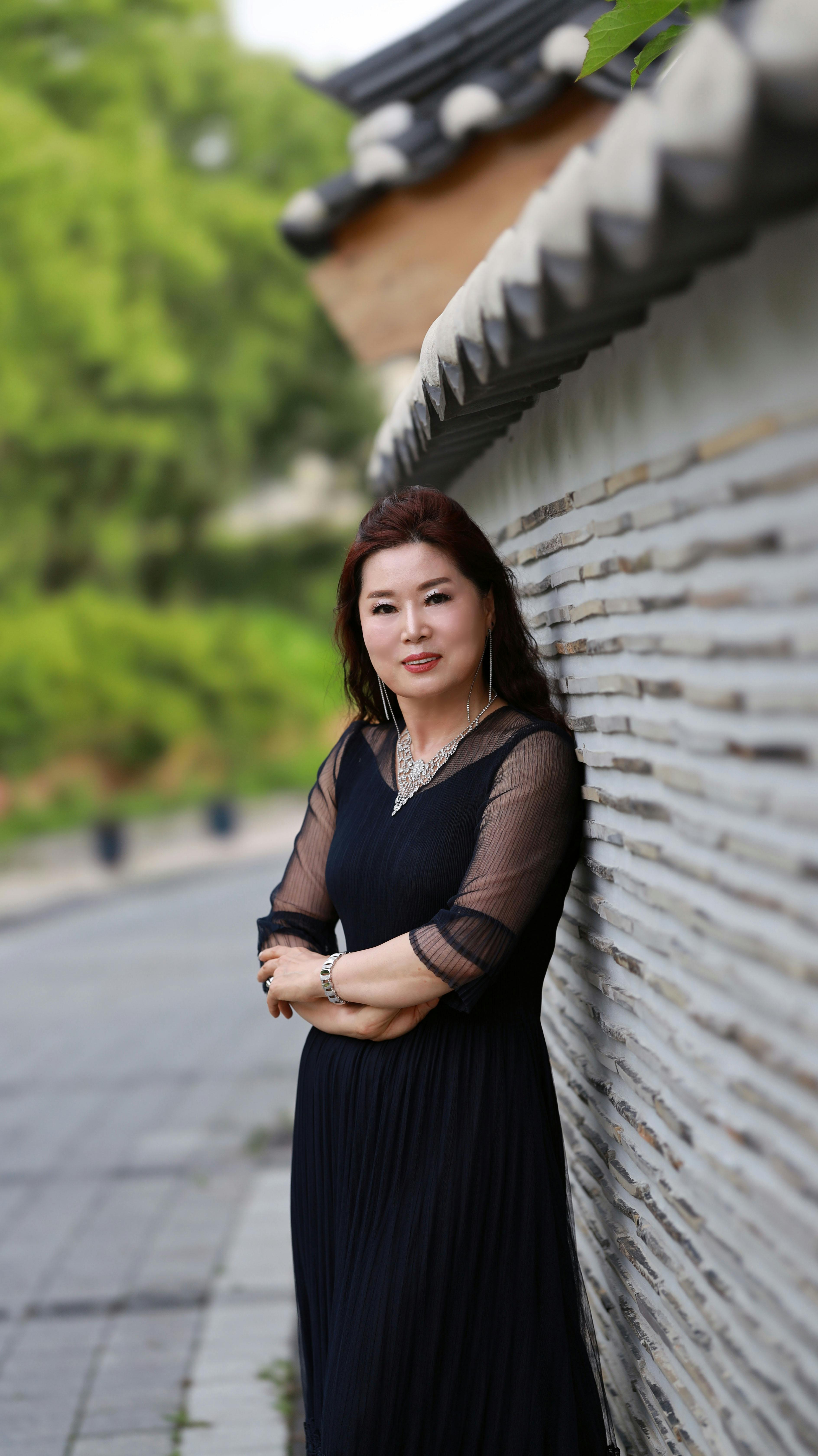Woman in a black dress smiling while leaning against a wall outdoors in a serene setting.
