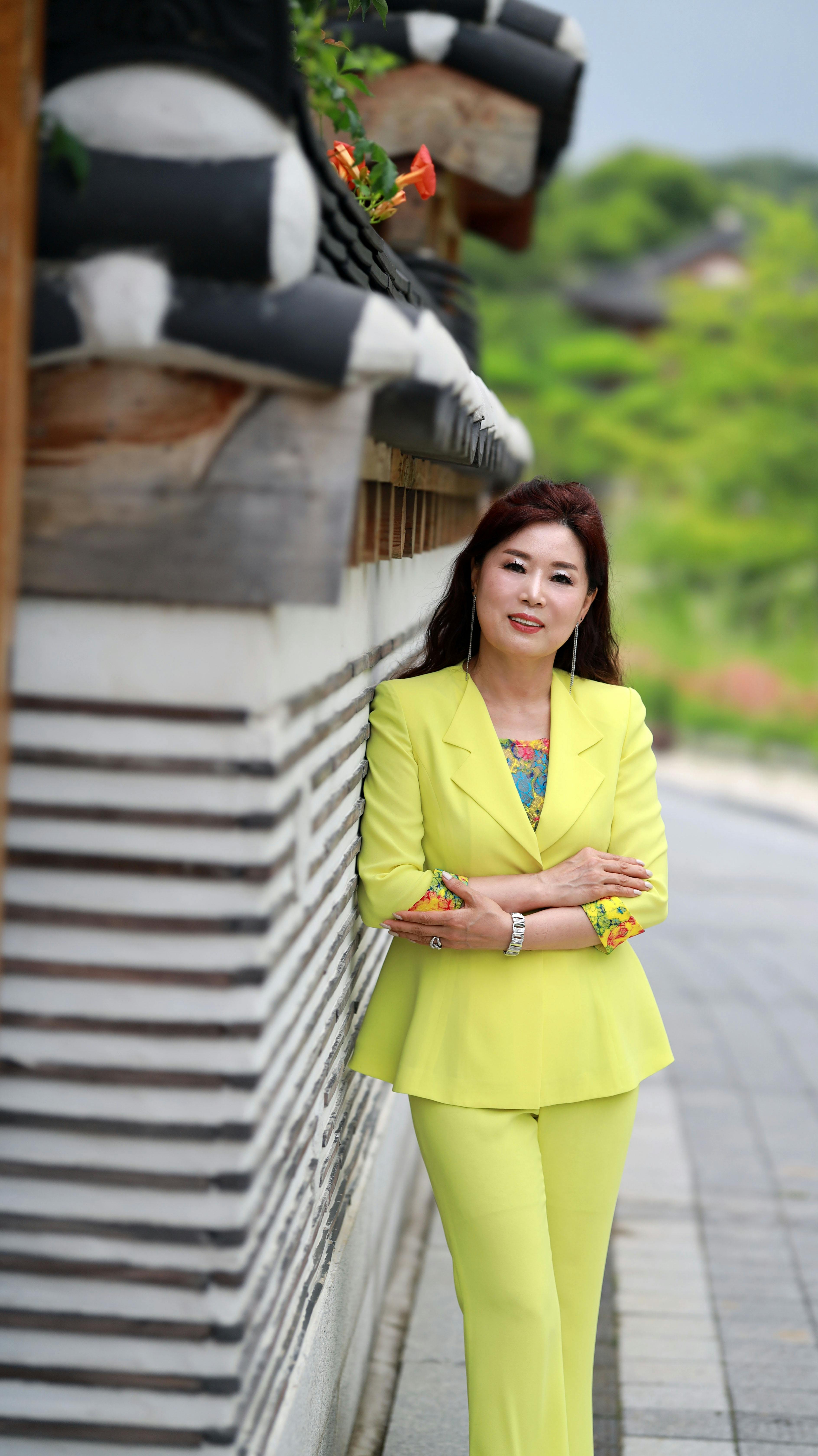 Asian woman in bright yellow suit stands confidently by an outdoor traditional wall.