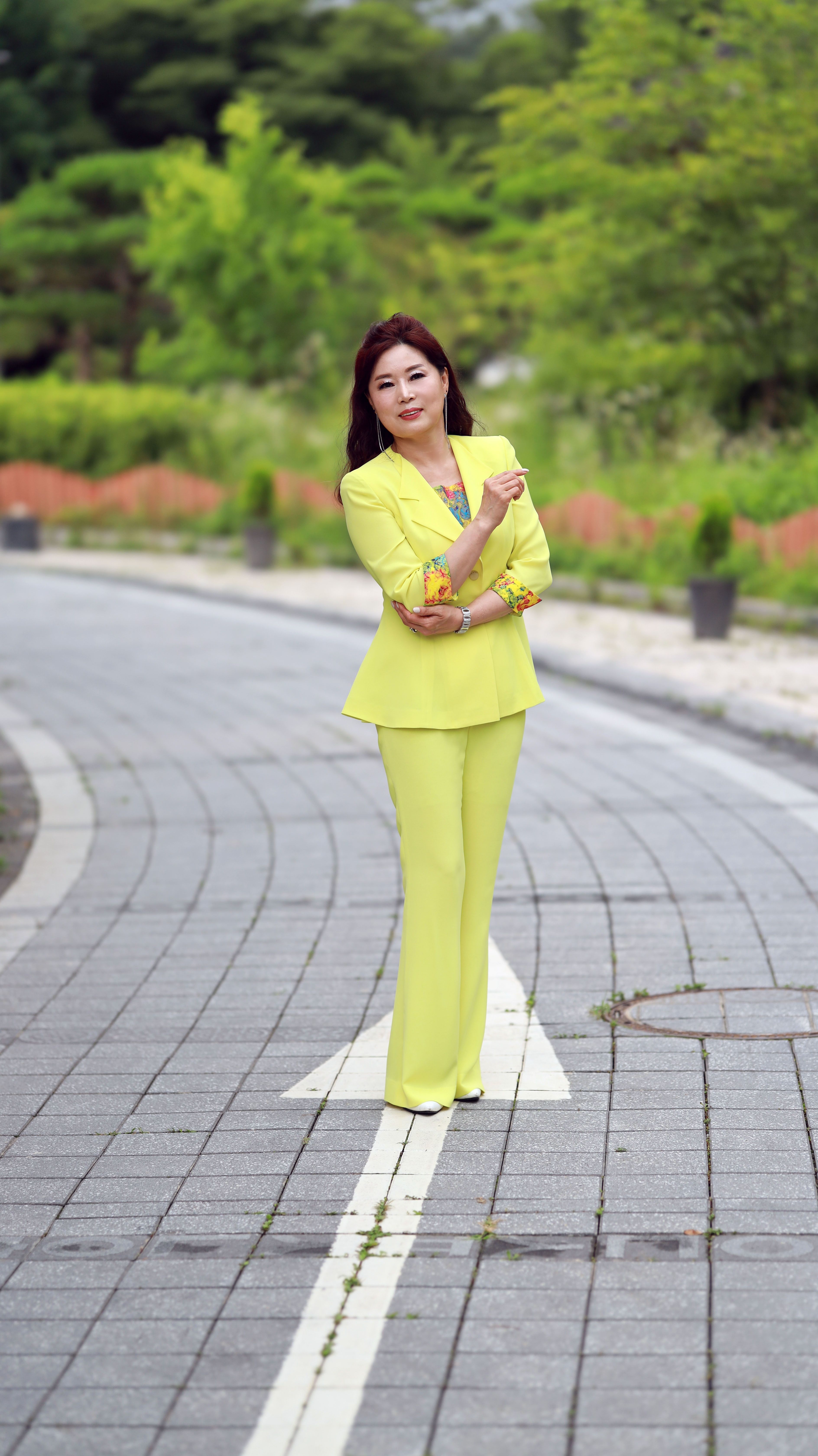 A woman in an elegant yellow suit stands confidently on a paved path in an outdoor setting.