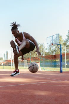 A young athletic man in activewear performs a dynamic basketball trick on an outdoor court.