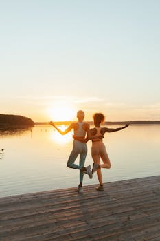 Two women in activewear perform yoga on a lakeside dock at sunrise for a serene workout.