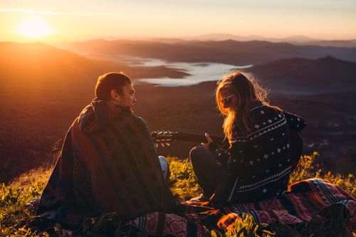 Free Couple enjoying a romantic sunset on a mountain peak while playing guitar, surrounded by nature. Stock Photo