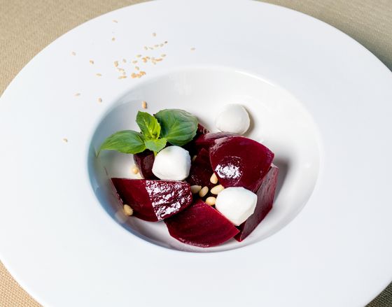 Roasted beet wedges with small mozzarella balls, basil sprig and pine nuts in white bowl