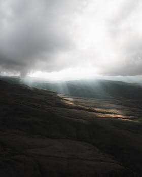 Sun rays piercing through a thick cloud cover illuminate a dramatic landscape, creating an awe-inspiring natural scene.
