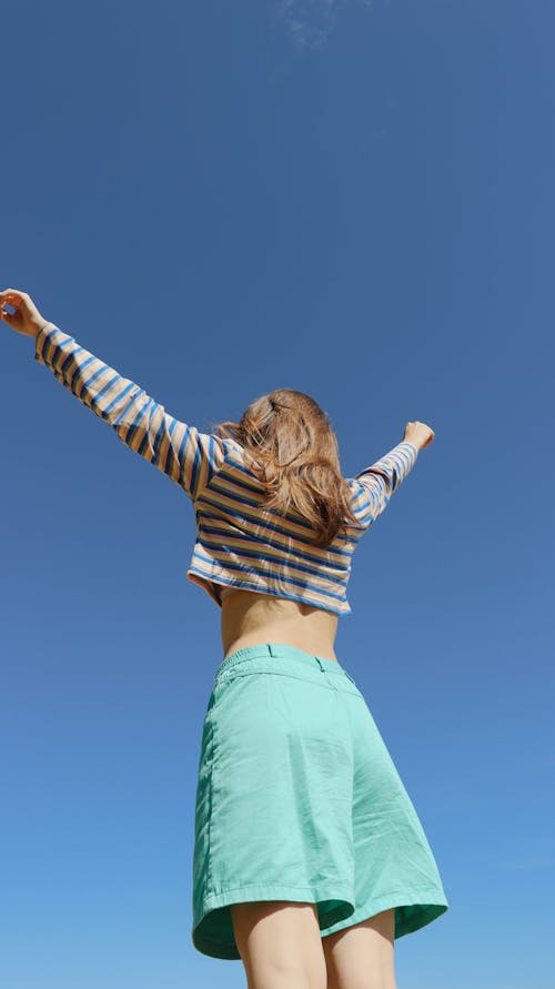 Woman in Striped Shirt and Green Shorts with Arms Raised Under Blue Sky