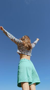 A young woman raises her arms in joy, wearing a striped shirt and green shorts against a clear blue sky.